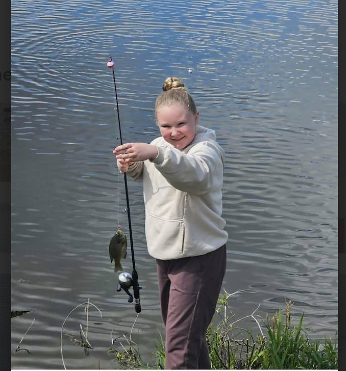 Student smiles while holding a fishing rod and a small fish beside a lake on a sunny day.