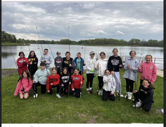 Students stand along a lakeshore holding fishing rods during an outdoor fishing trip under cloudy skies.