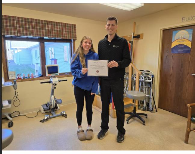 Student and staff member smile while holding a certificate inside a rehabilitation or therapy room with exercise equipment nearby.