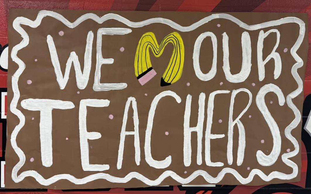 Hand-painted sign reads “We *Heart* Our Teachers,” with a yellow pencil forming  on a brown background.