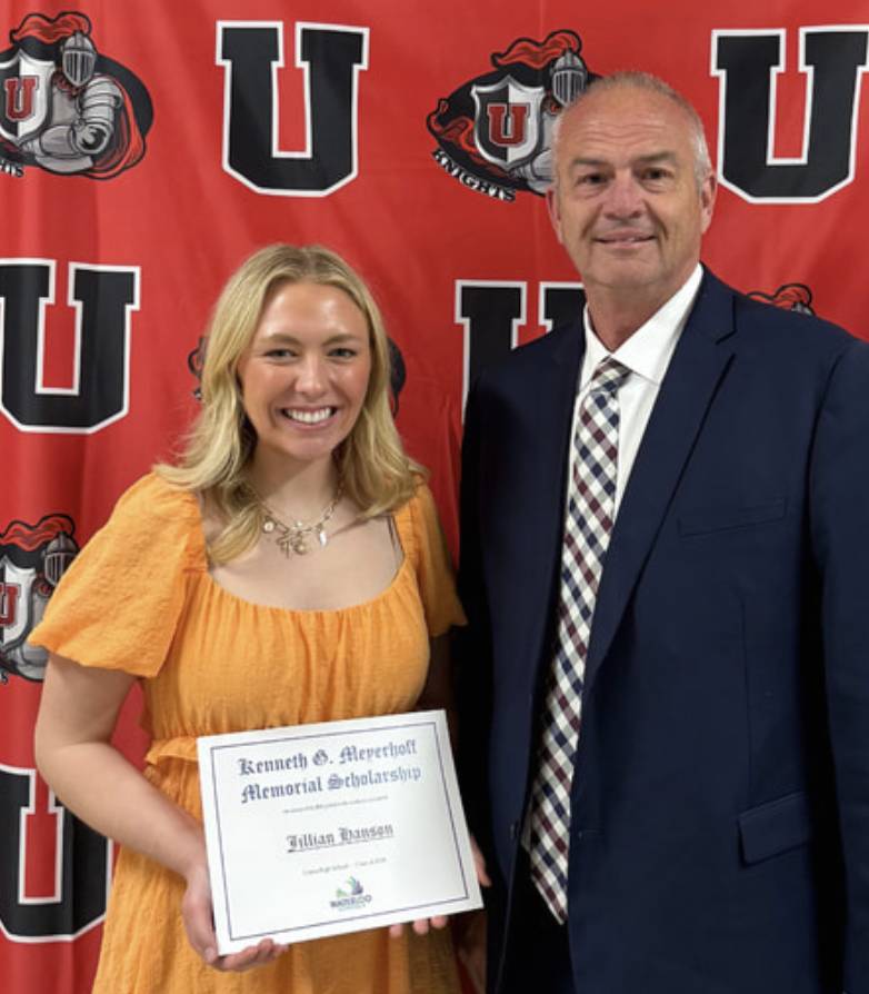 Smiling scholarship recipient in a yellow dress holds certificate beside a school administrator at an awards event.