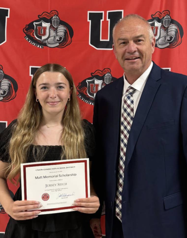 Scholarship recipient poses with certificate beside a school administrator in front of a red Union Knights backdrop.