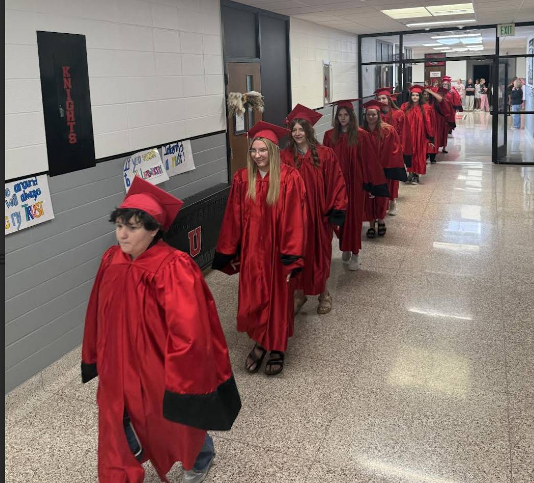 Graduating seniors in red caps and gowns walk through a school hallway in a graduation procession.