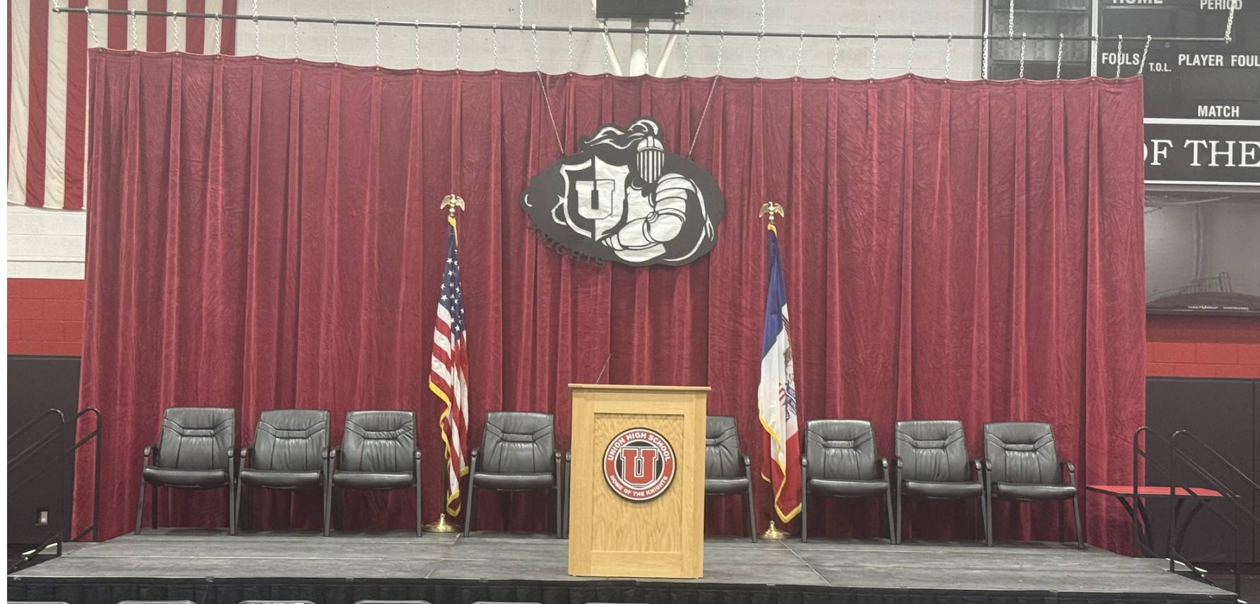 Graduation stage with red curtains, podium, flags, and chairs set up inside a school gymnasium.