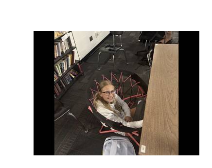 Student smiles while sitting in a webbed lounge chair beside a classroom desk and bookshelf.