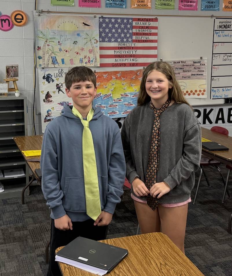 Two students smile in a classroom while wearing oversized neckties during a fun dress-up activity.