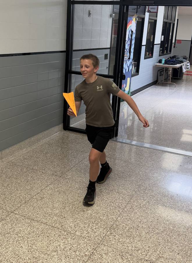 Student walks through a school hallway holding a bright orange paper airplane during a classroom activity.