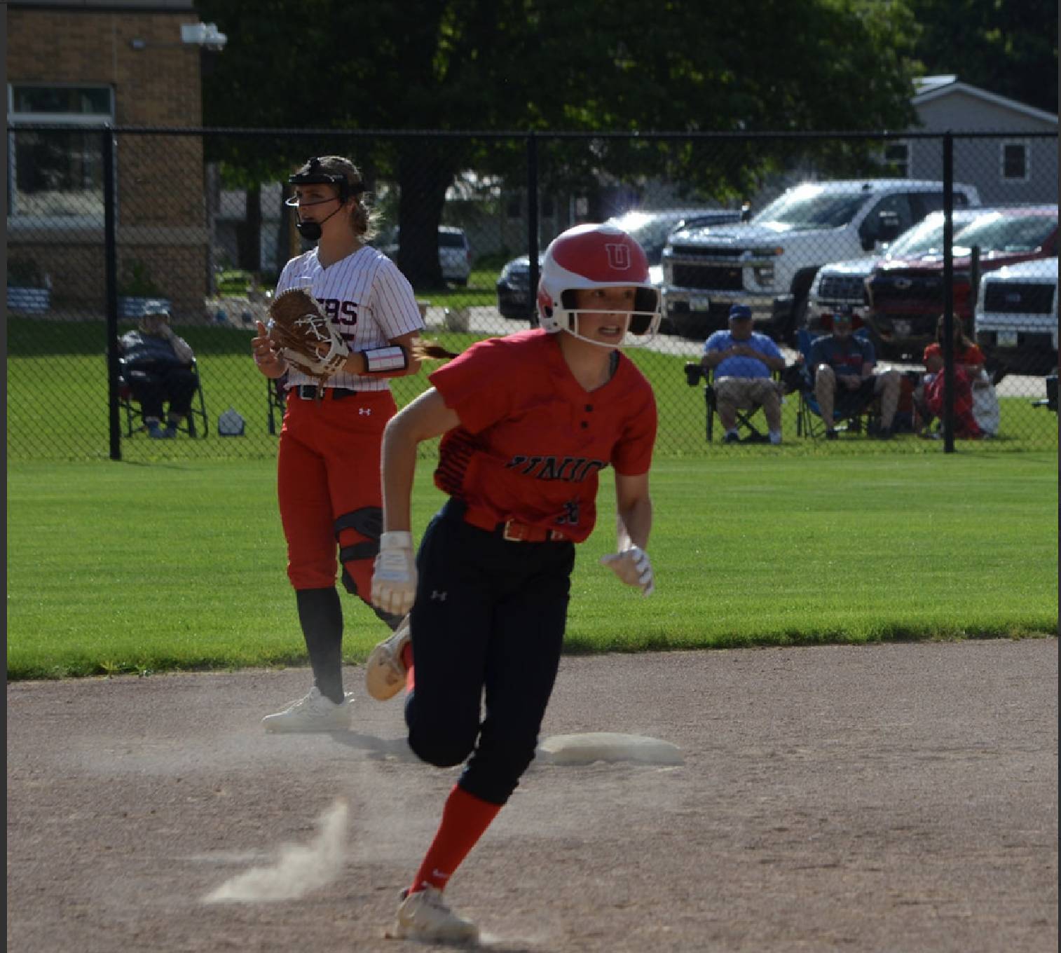 Softball player rounds a base at full speed, kicking up dust while a fielder watches from the infield during a game.
