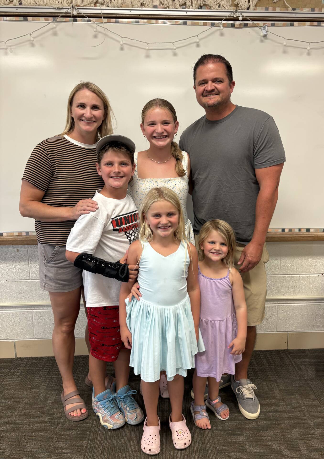 Two adults and three smiling children pose together in a classroom, gathered closely in front of a whiteboard.