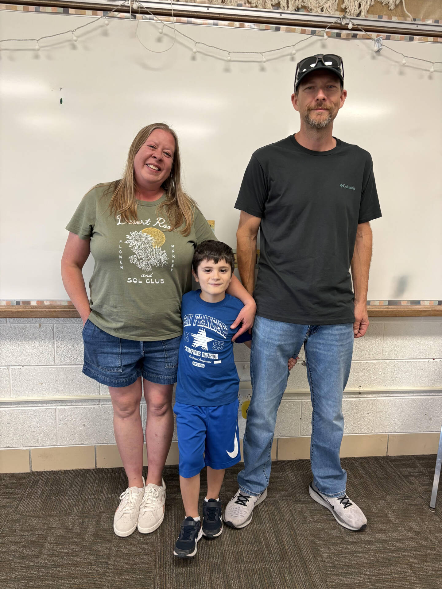 Two adults stand with a smiling young boy in blue athletic clothes, posing together in front of a classroom whiteboard.