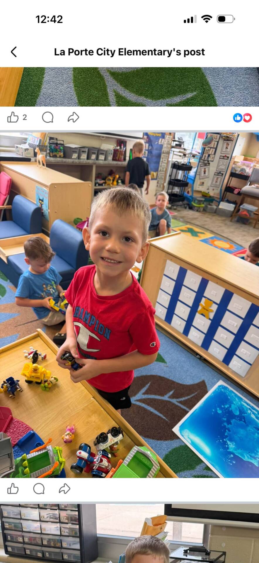 A smiling student holds small toys at a classroom activity table while classmates play nearby.
