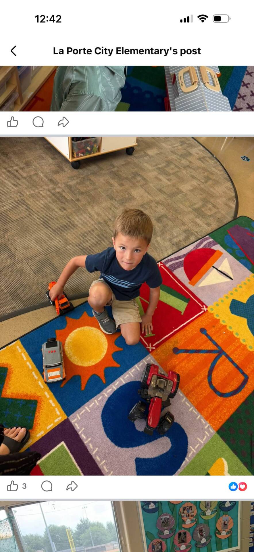 A student kneels on a colorful classroom rug while playing with toy trucks and a tractor.