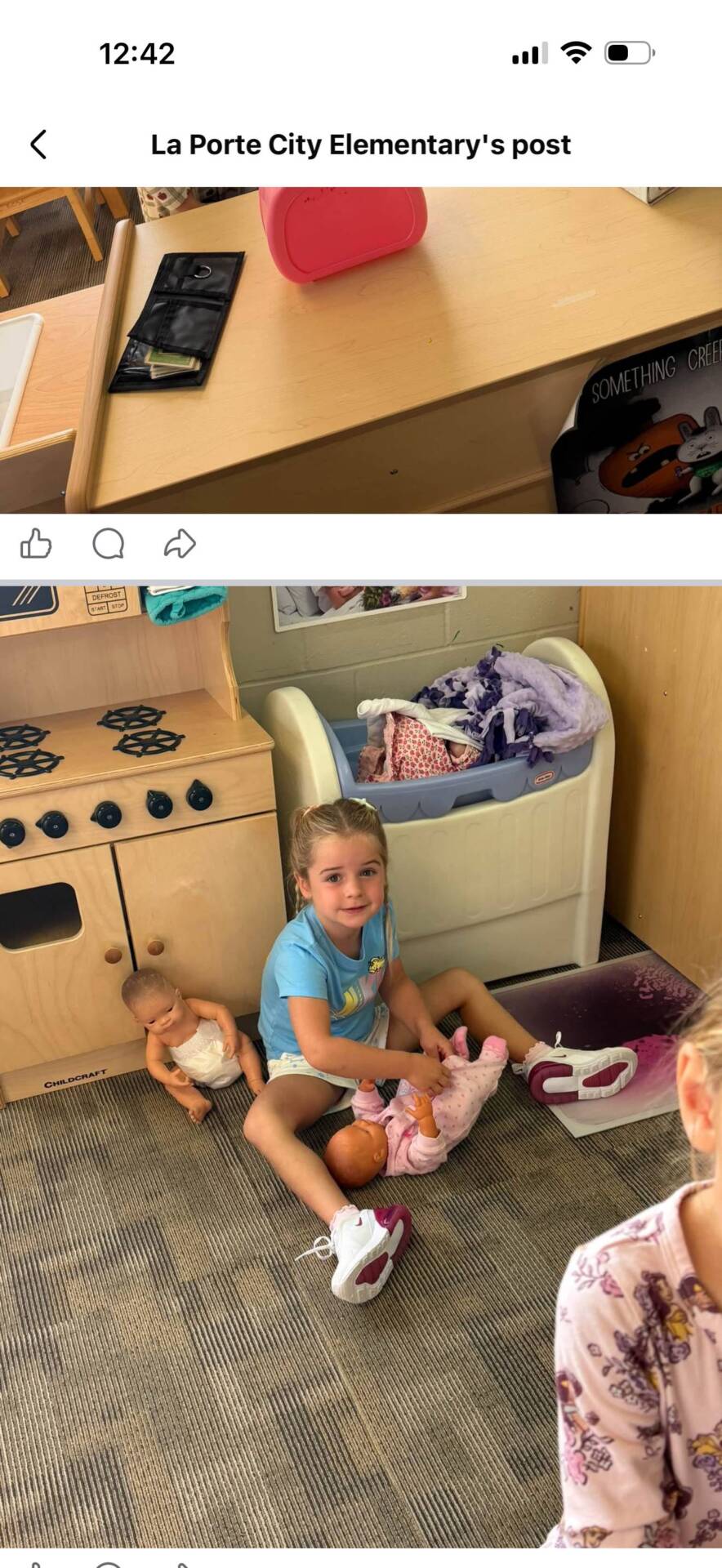 A student sits in a classroom play area caring for baby dolls beside a toy kitchen and doll bed.