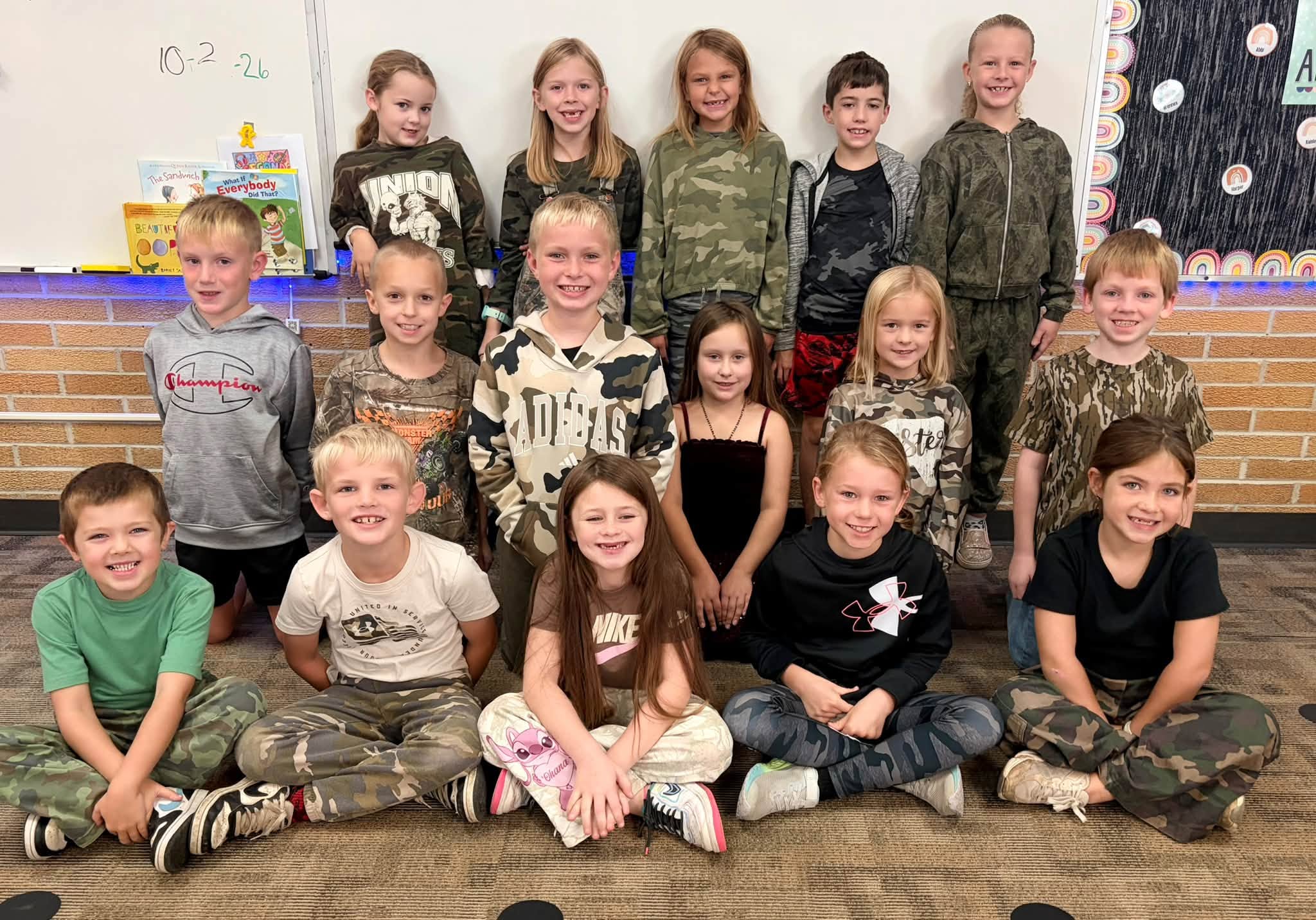 Fifteen elementary students wearing camouflage and casual clothing pose together in three rows in front of a classroom whiteboard.