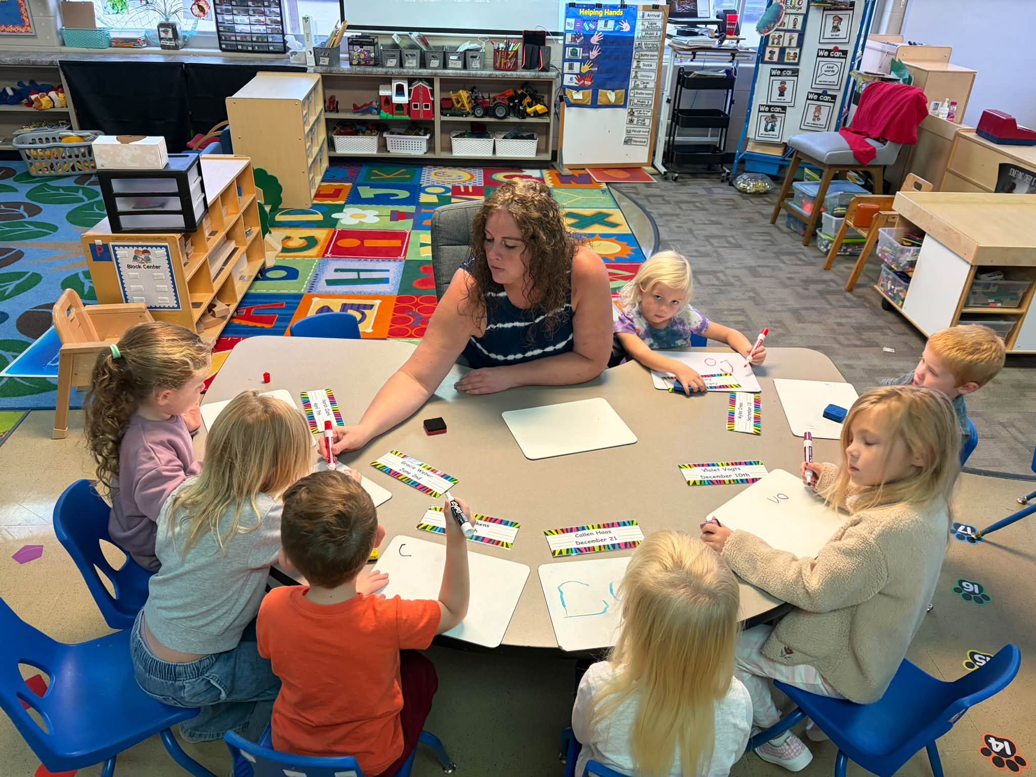 A preschool teacher helps young students practice writing on whiteboards around a classroom table.