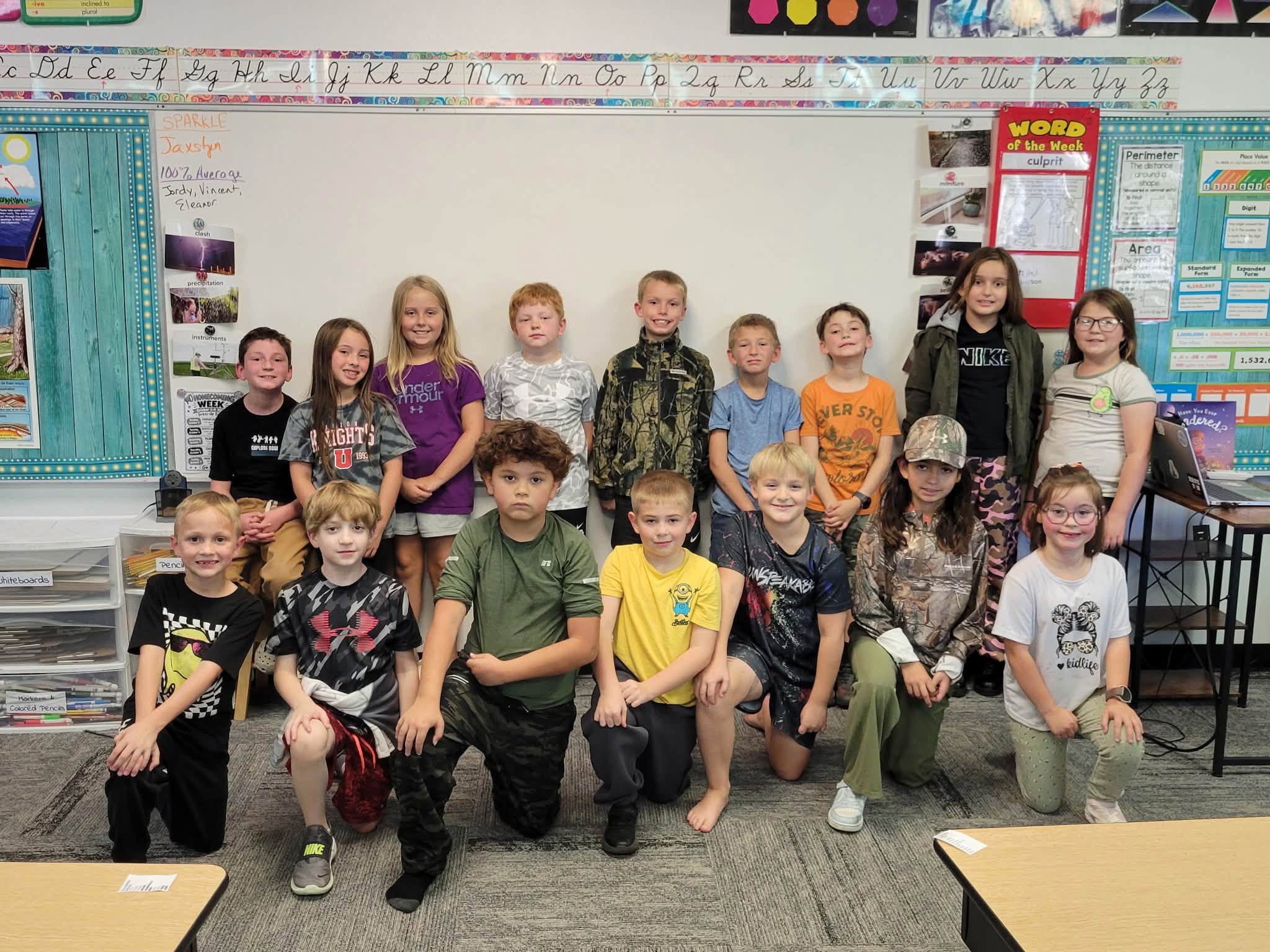Fifteen elementary students pose together in two rows in a classroom, smiling in front of a whiteboard.