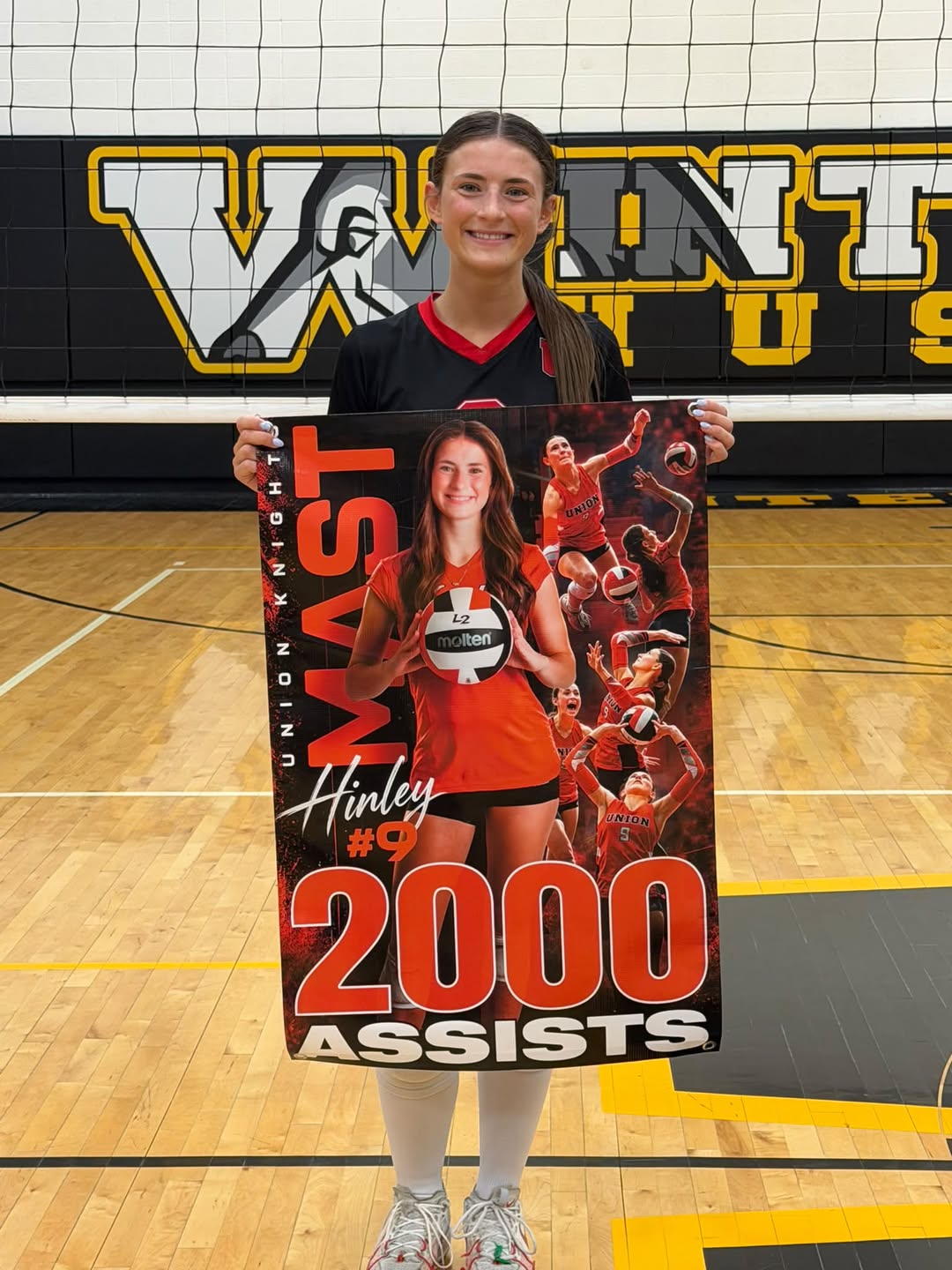 A smiling volleyball player holds a poster celebrating her milestone of 2,000 career assists in a school gymnasium.
