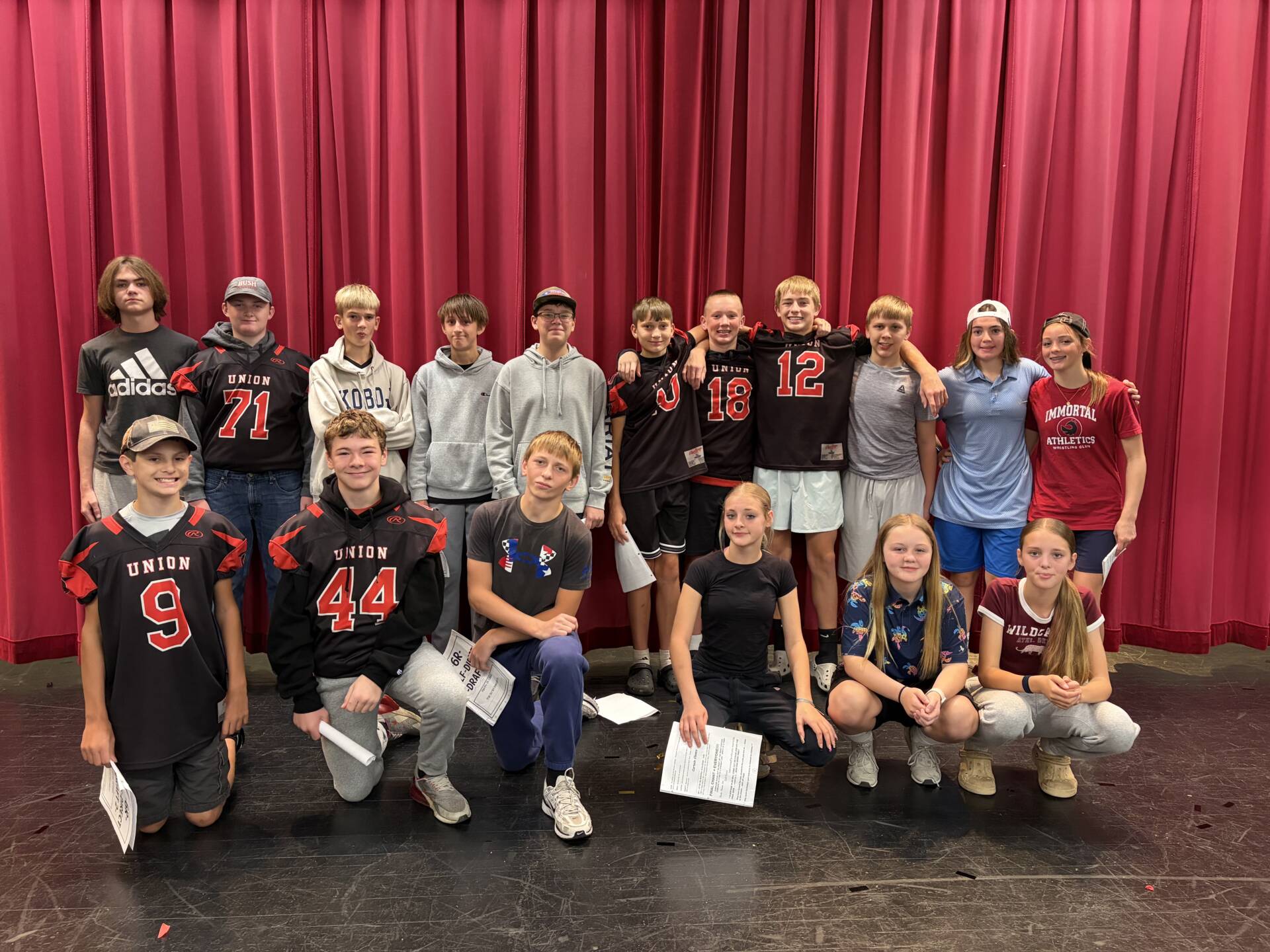 Seventeen middle school students, some wearing Union football jerseys, pose together onstage in front of red curtains.