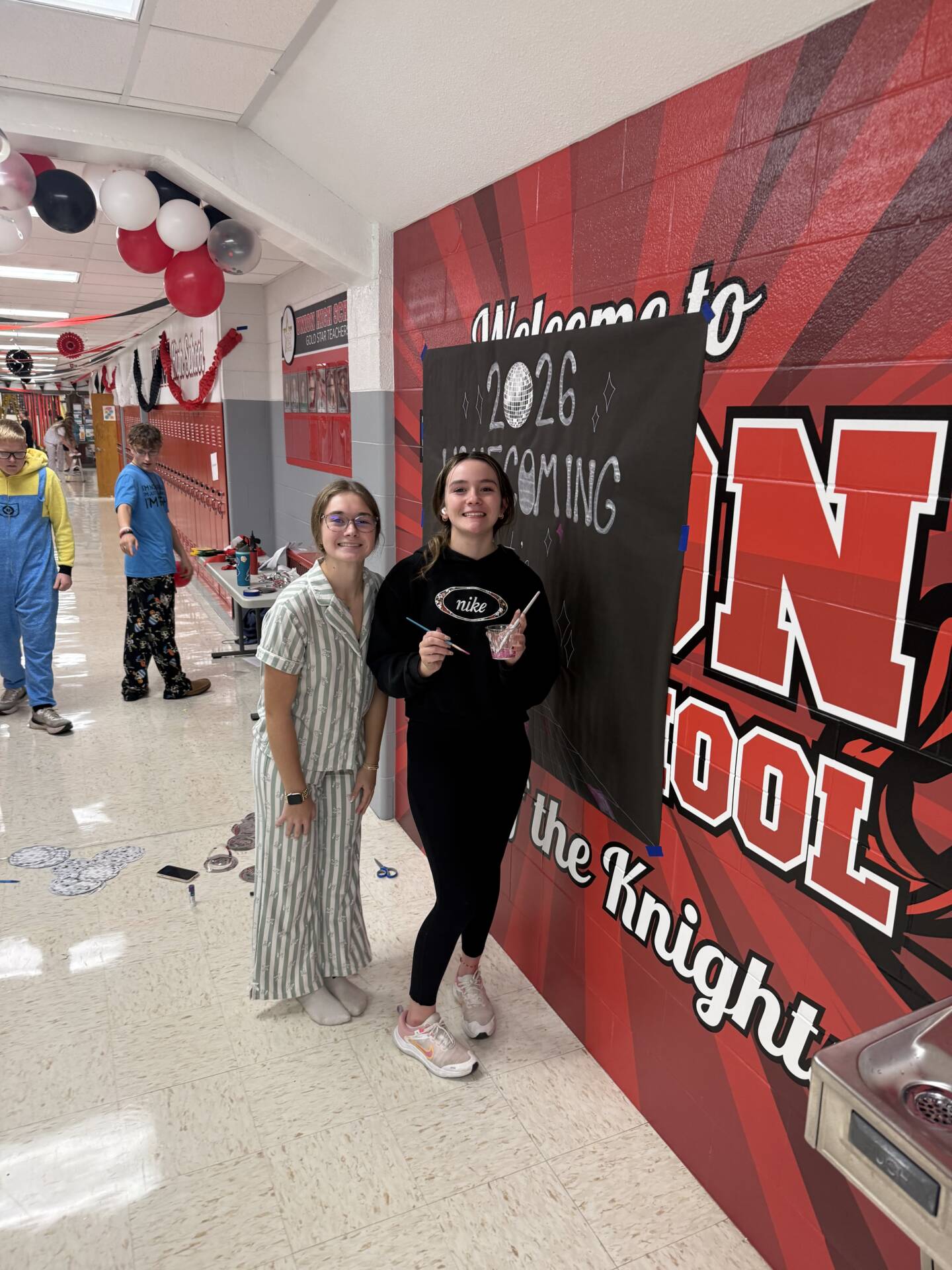Students decorate a school hallway for homecoming with a black 2026 Homecoming sign and red, black, and white balloons.