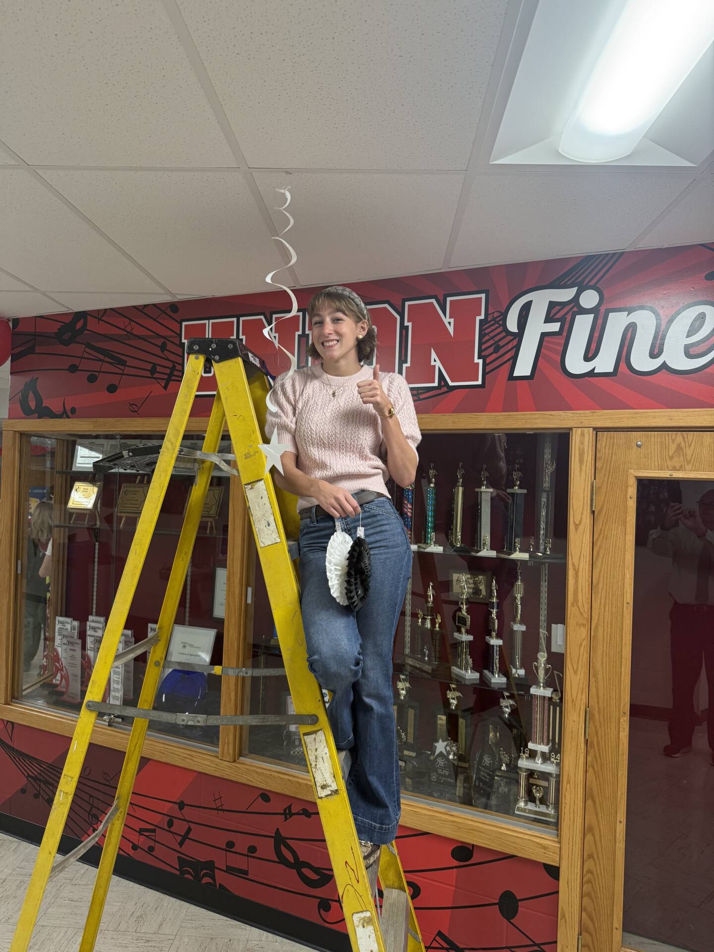 A smiling student stands on a yellow ladder, giving a thumbs-up while hanging decorations near a school trophy case.