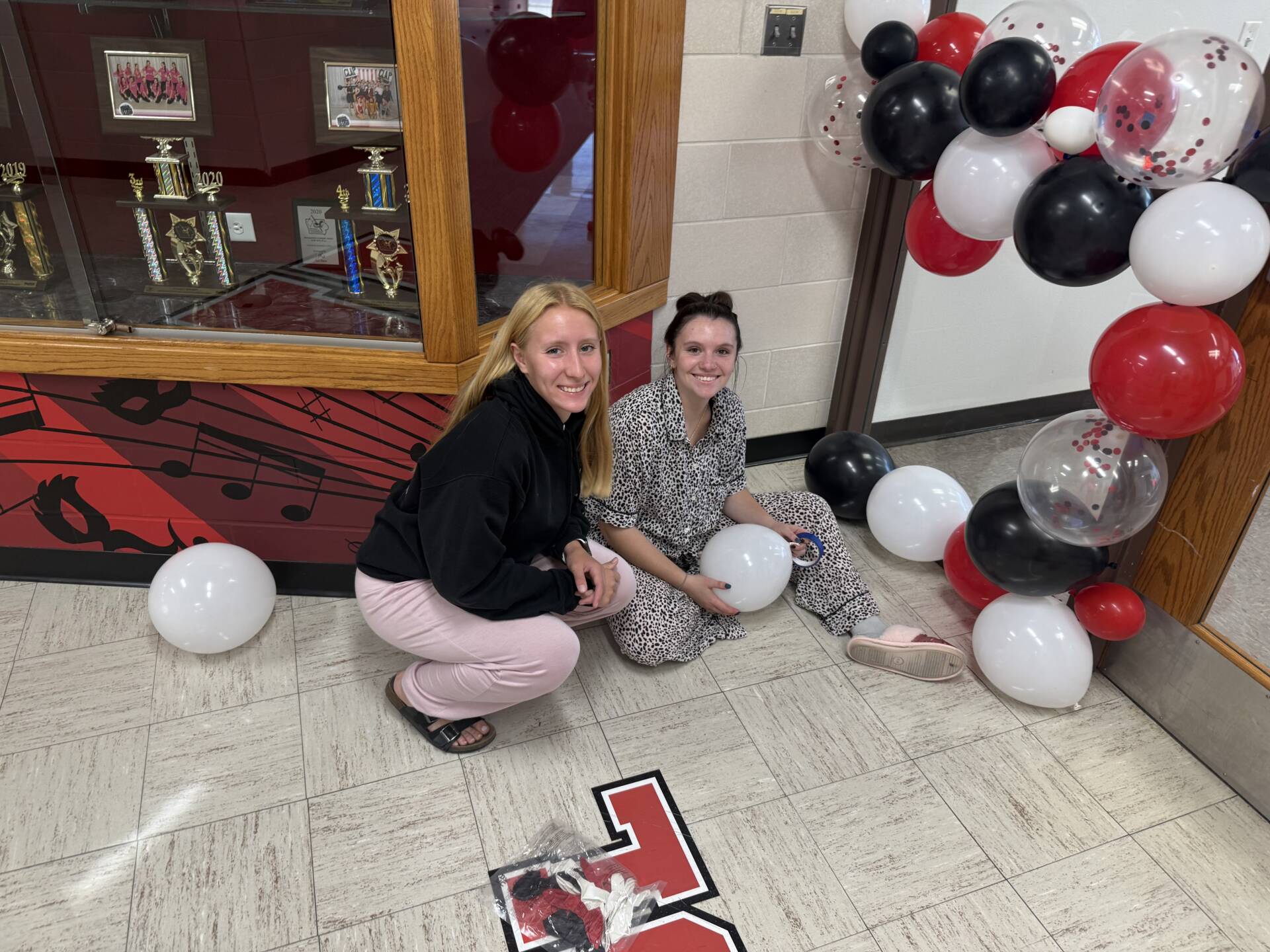 Two women smile while assembling a red, black, and white balloon display in a school hallway.
