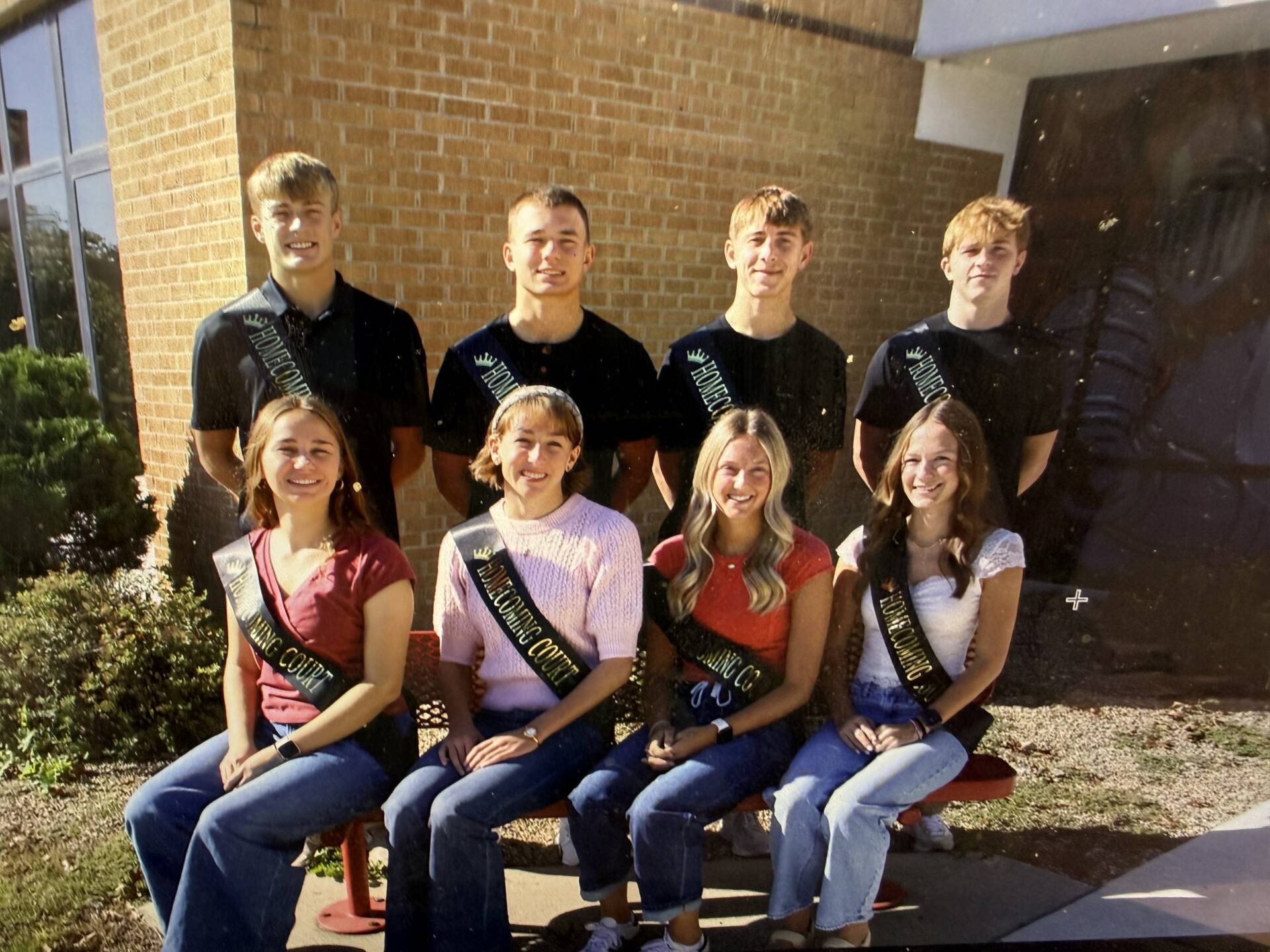 Eight homecoming court members wearing sashes pose outdoors in two rows in front of a brick school building.
