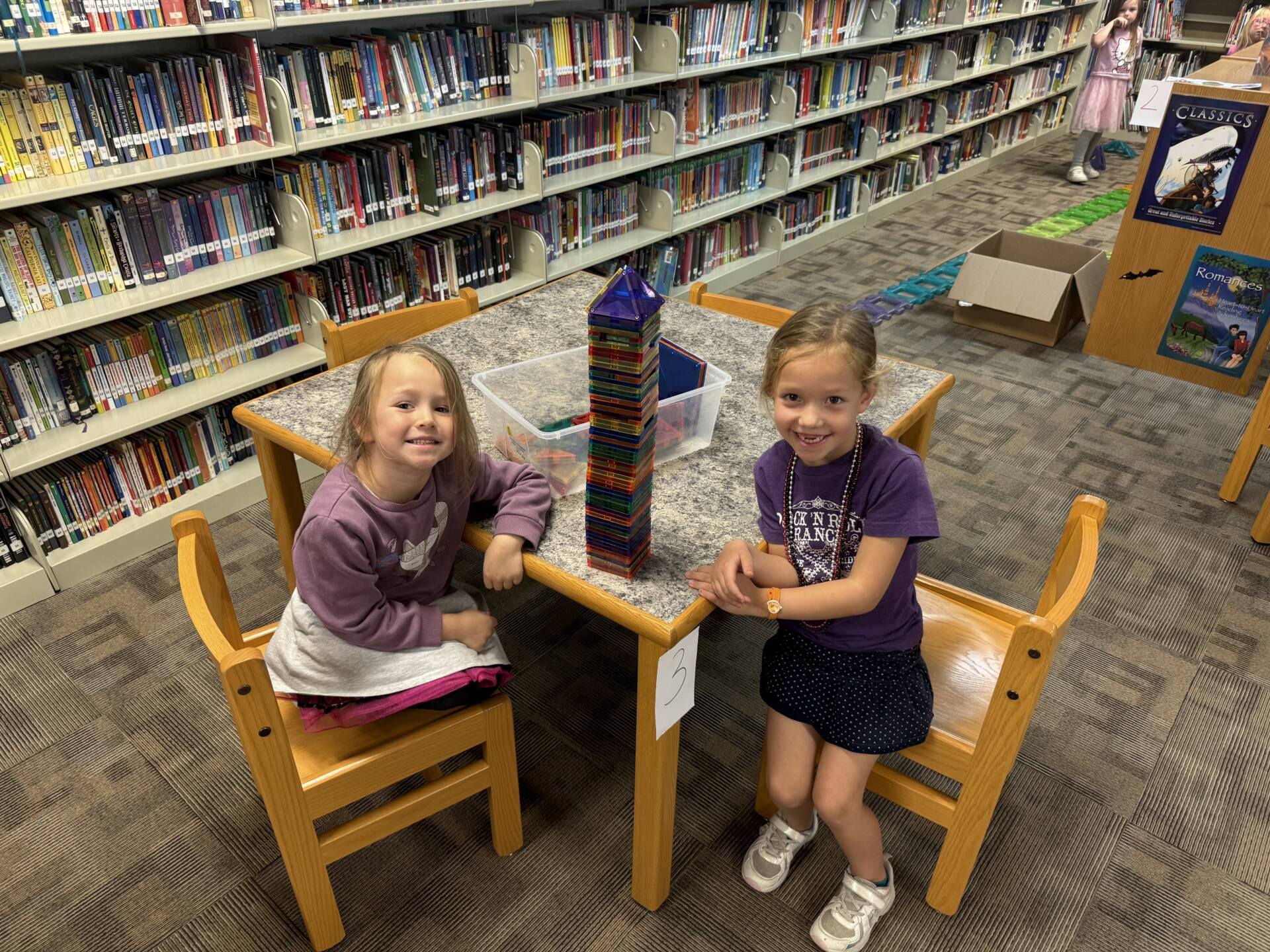 Two smiling young girls sit beside a tall, colorful magnetic tile tower they built together in a school library.