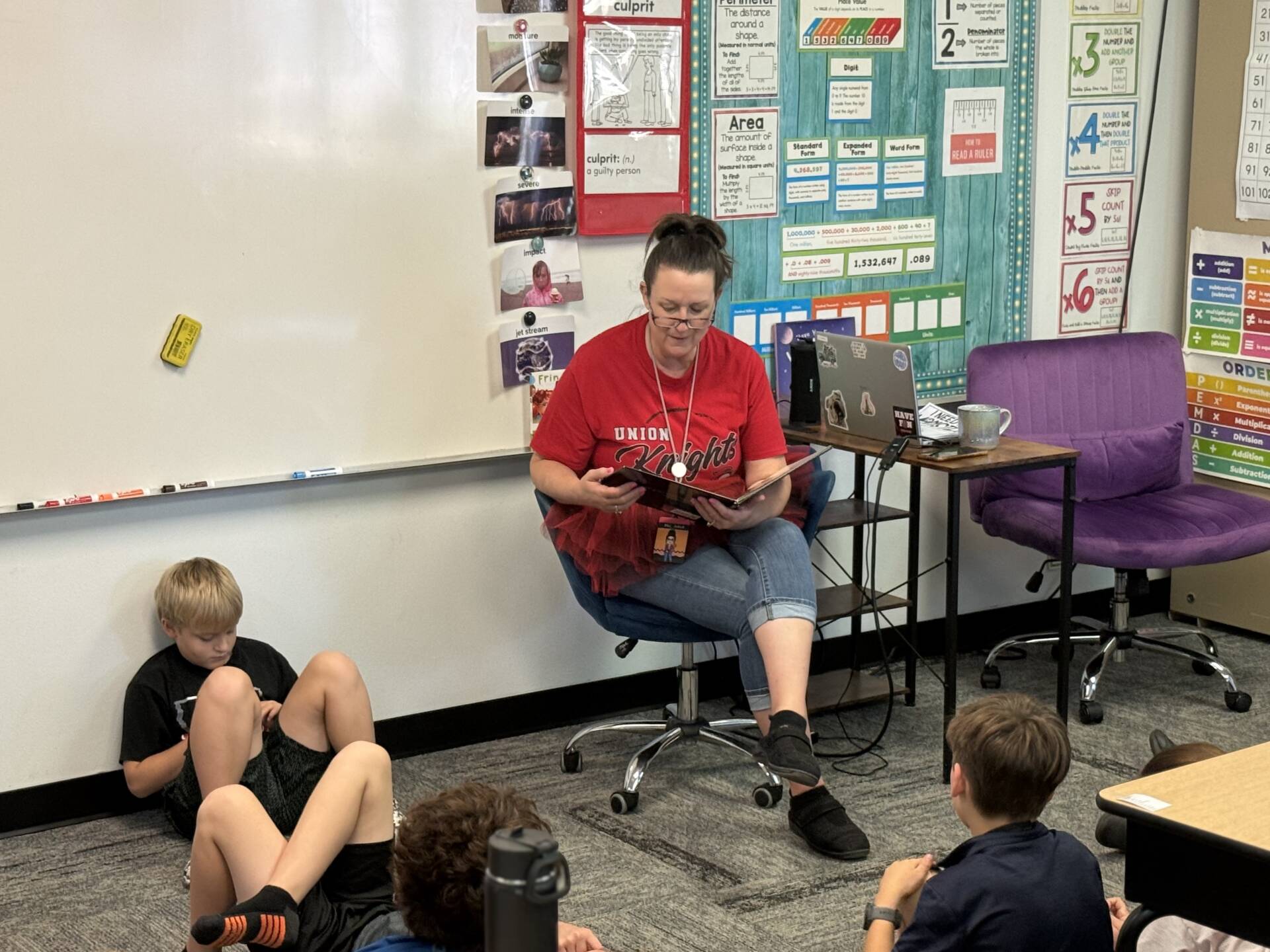A teacher in a red Union Knights shirt reads aloud to students seated on the classroom floor.