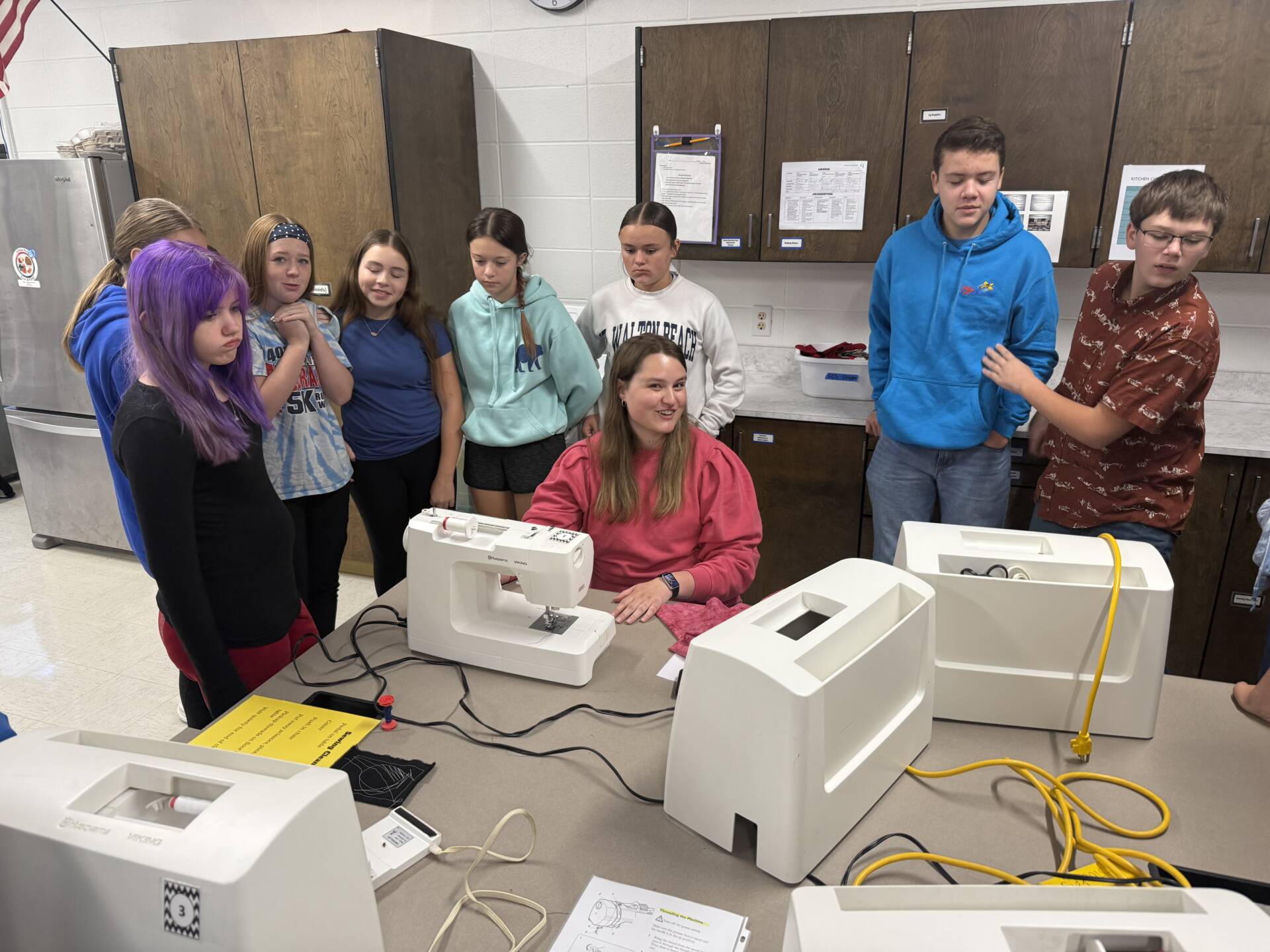 A teacher in a pink sweatshirt demonstrates a sewing machine to students gathered around a table in a classroom.