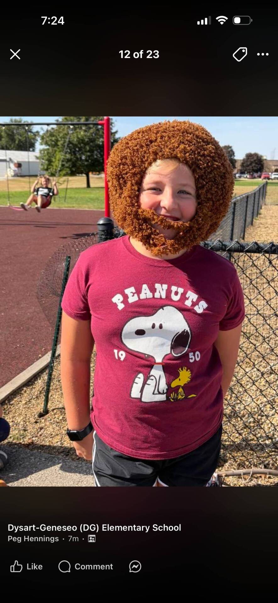 Student wearing a curly brown wig and beard smiles outdoors in a maroon Peanuts T-shirt near a playground.
