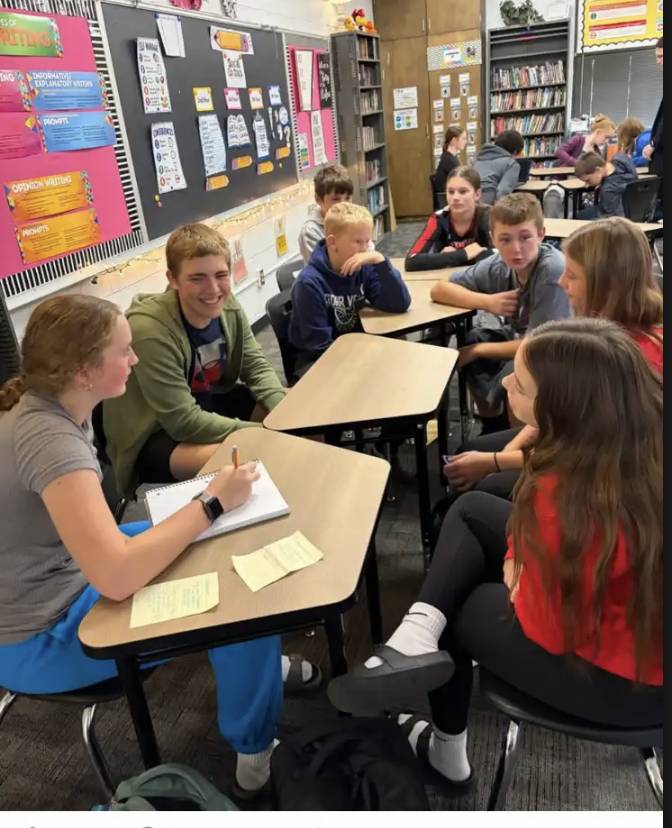 Students sit in small groups at classroom desks, smiling and discussing assignments with notebooks and papers.