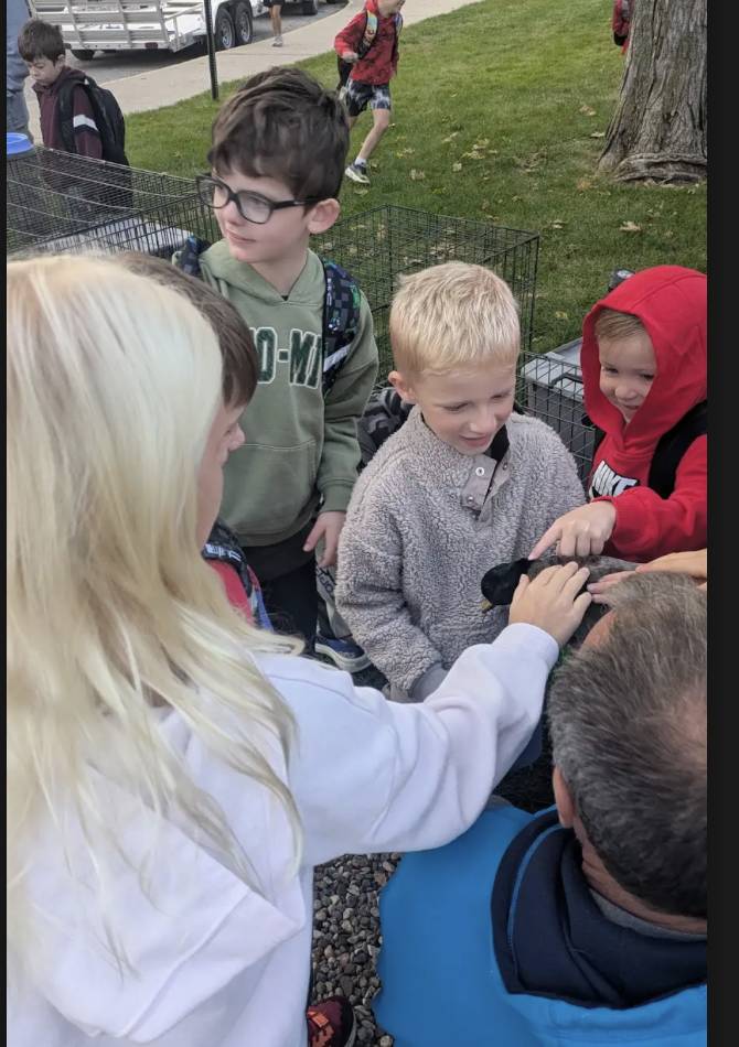 Students gather around a small black animal while an adult helps them gently pet it outside near animal cages.