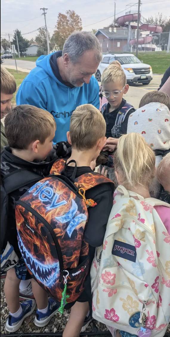 A man in a blue hoodie gathers with elementary students wearing backpacks as they examine something together outdoors.