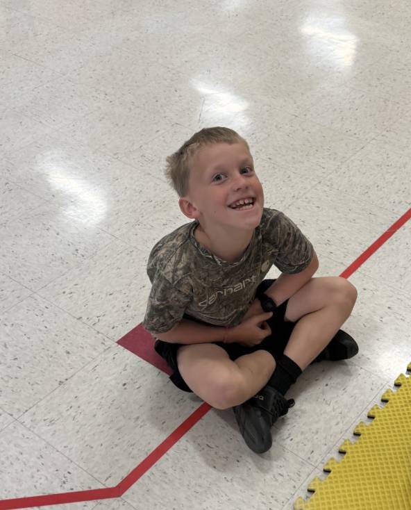 A smiling young boy in a camouflage shirt sits cross-legged on a classroom floor beside colorful foam mats.