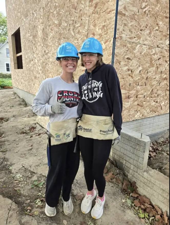 Two smiling volunteers wearing blue hard hats and Habitat for Humanity tool belts pose at a home construction site.