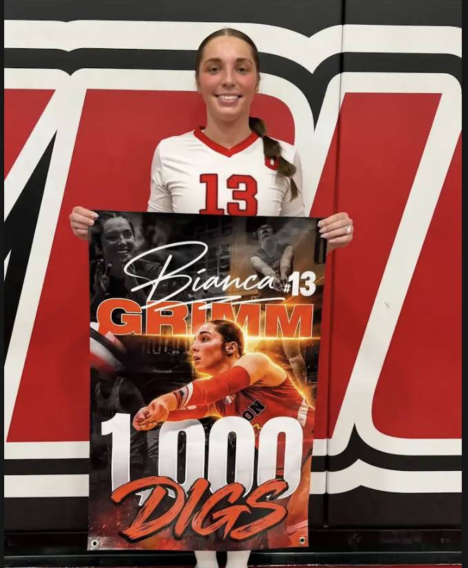 Union volleyball player Bianca Grimm, wearing jersey number 13, holds a banner celebrating her milestone of 1,000 career digs.