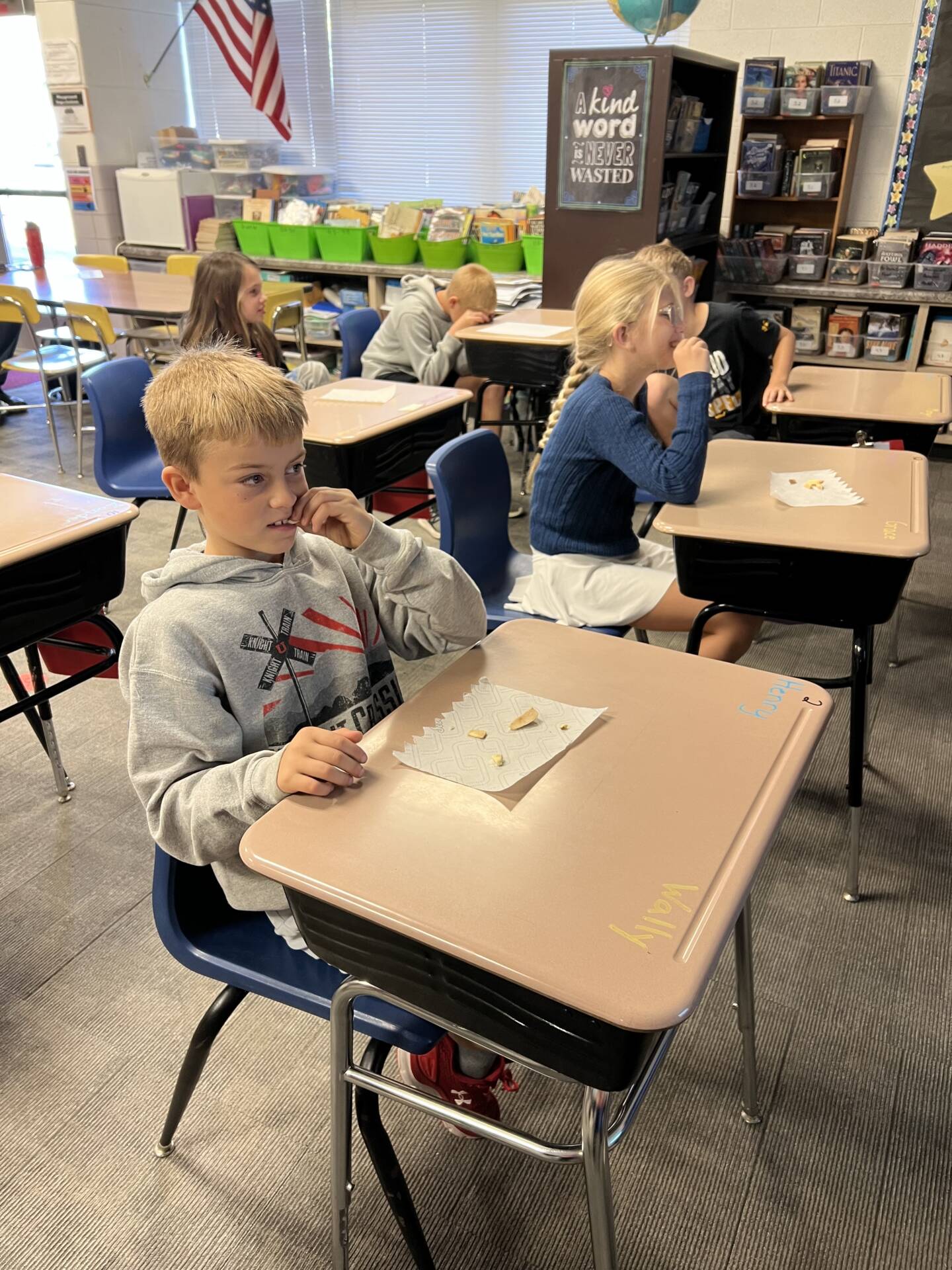 Elementary students sit at individual classroom desks while tasting snacks and interacting with their classmates.