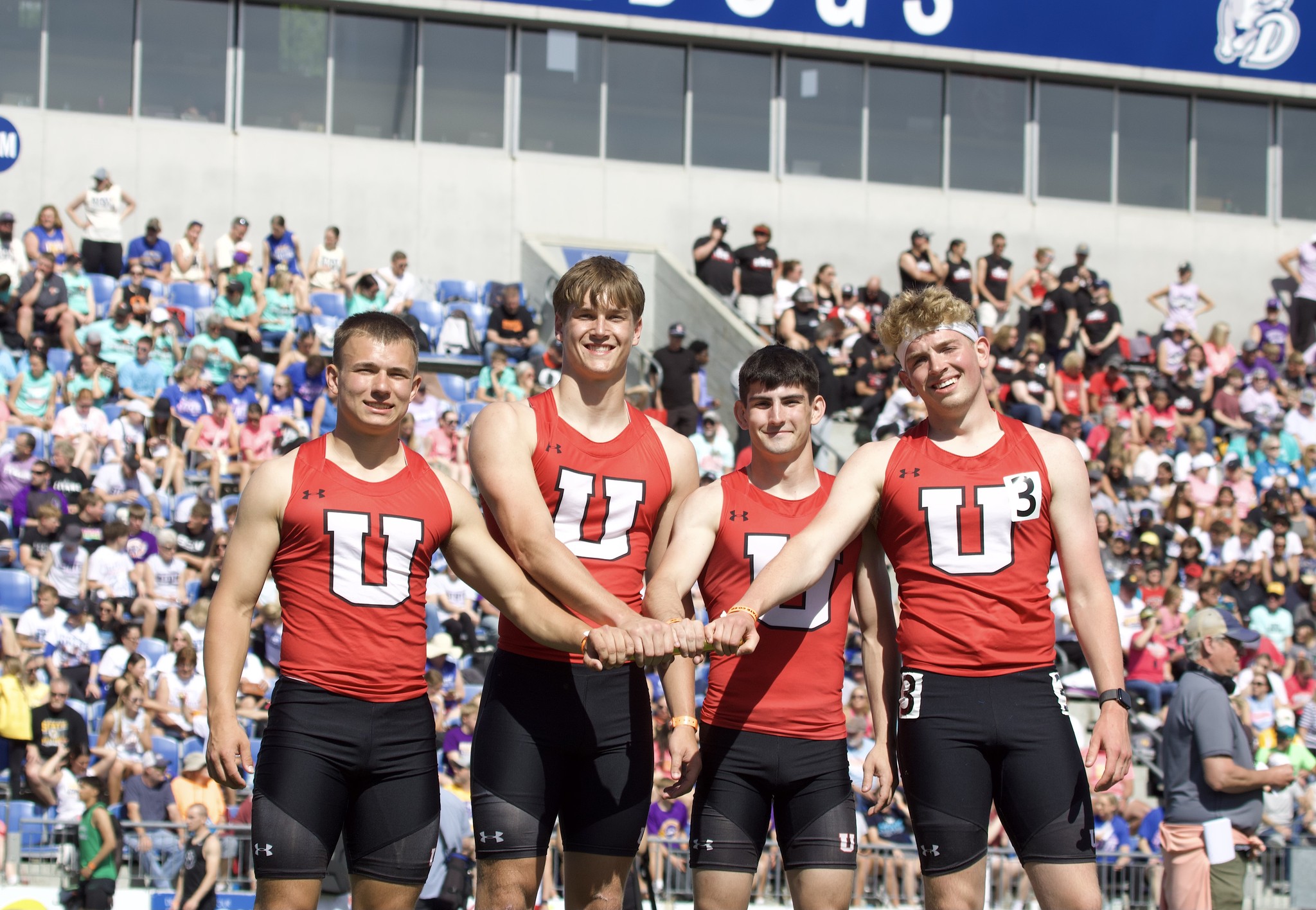 Four boys’ relay team members in red uniforms pose with a baton on the track at a state meet, smiling in front of a crowded stadium.