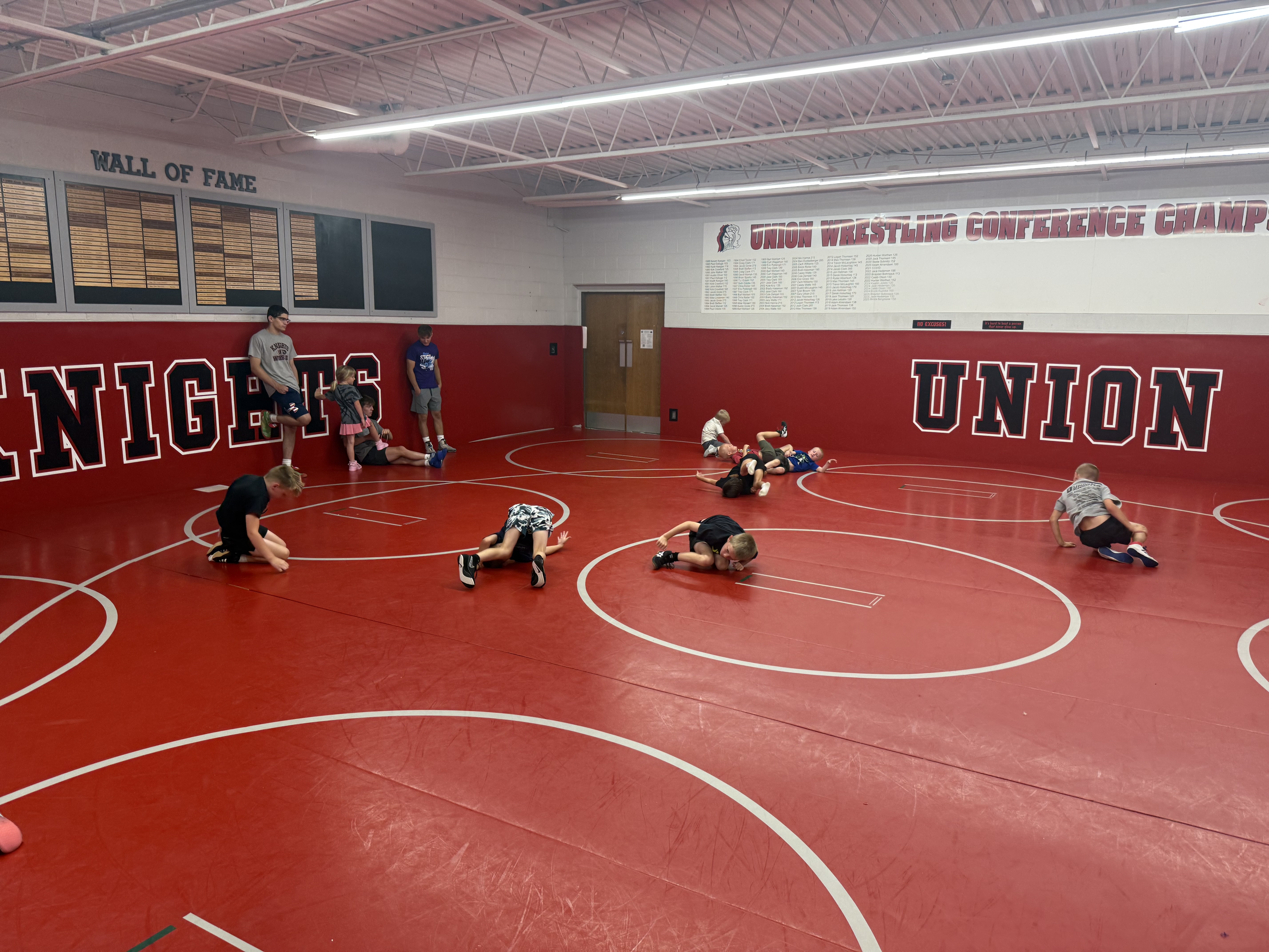 Young athletes practice wrestling drills on red mats in a high school wrestling room while coaches supervise from the sidelines.