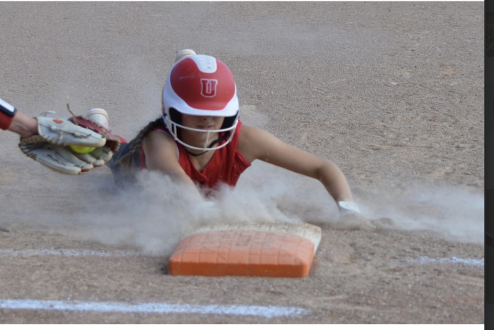 Softball player in a red uniform slides headfirst into a base, kicking up a cloud of dust as a fielder reaches in with the ball.